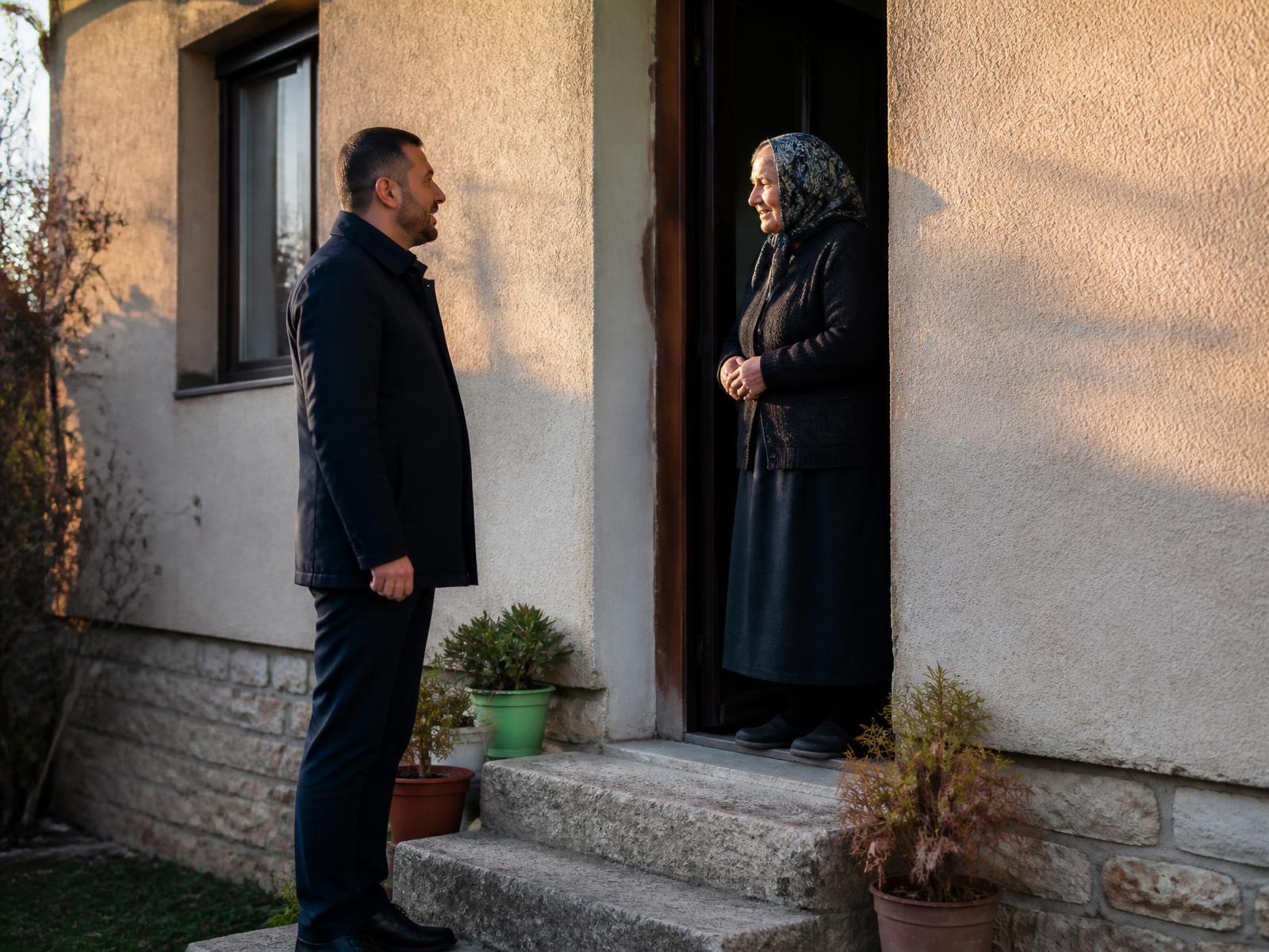 Muškarac u tamnoj jakni razgovara sa starijom ženom na kućnom pragu porodične kuće u popodnevnoj svjetlosti